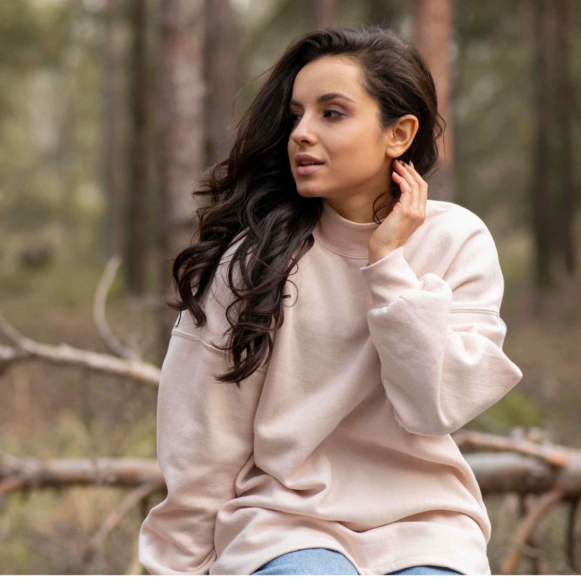 Woman with long dark hair sits in a forest wearing a cream sweater. She looks to the side, exuding a calm and thoughtful vibe.