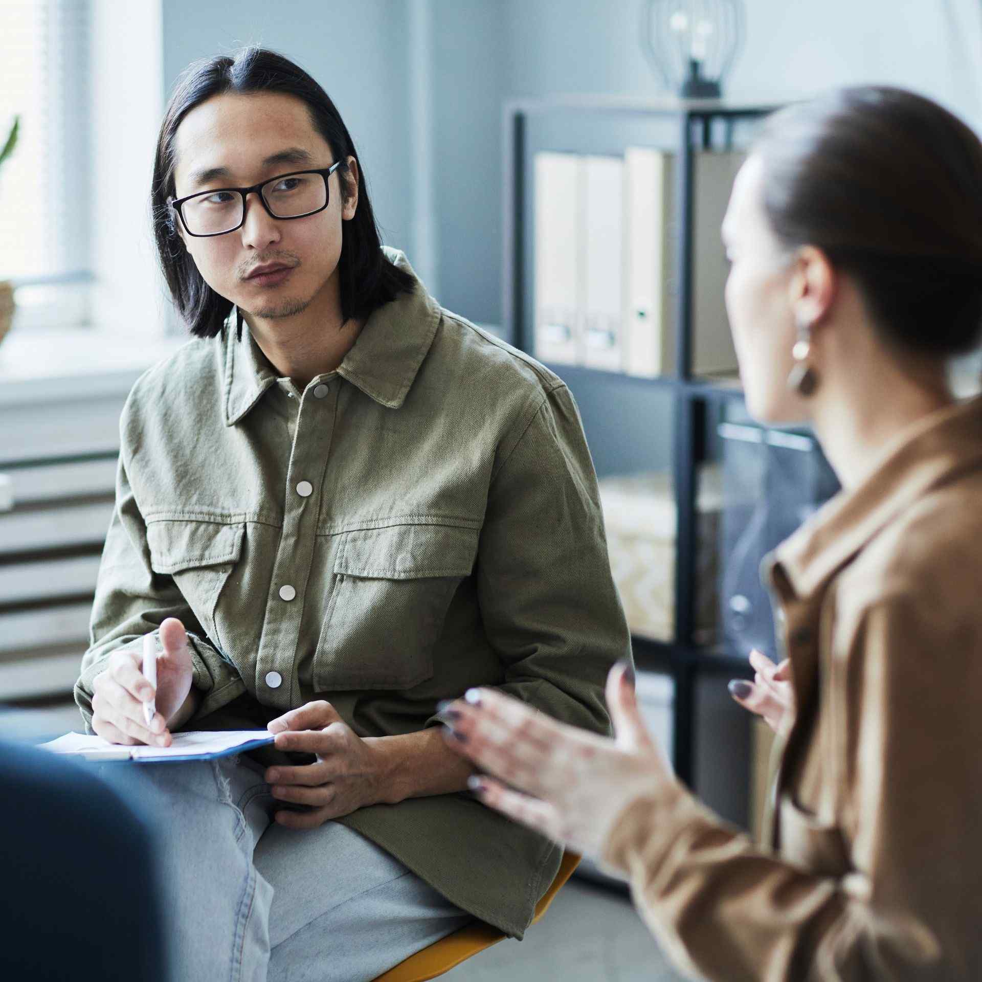 A man with glasses and long hair listens attentively to a woman gesturing during a discussion in a modern office setting, conveying focus and engagement.