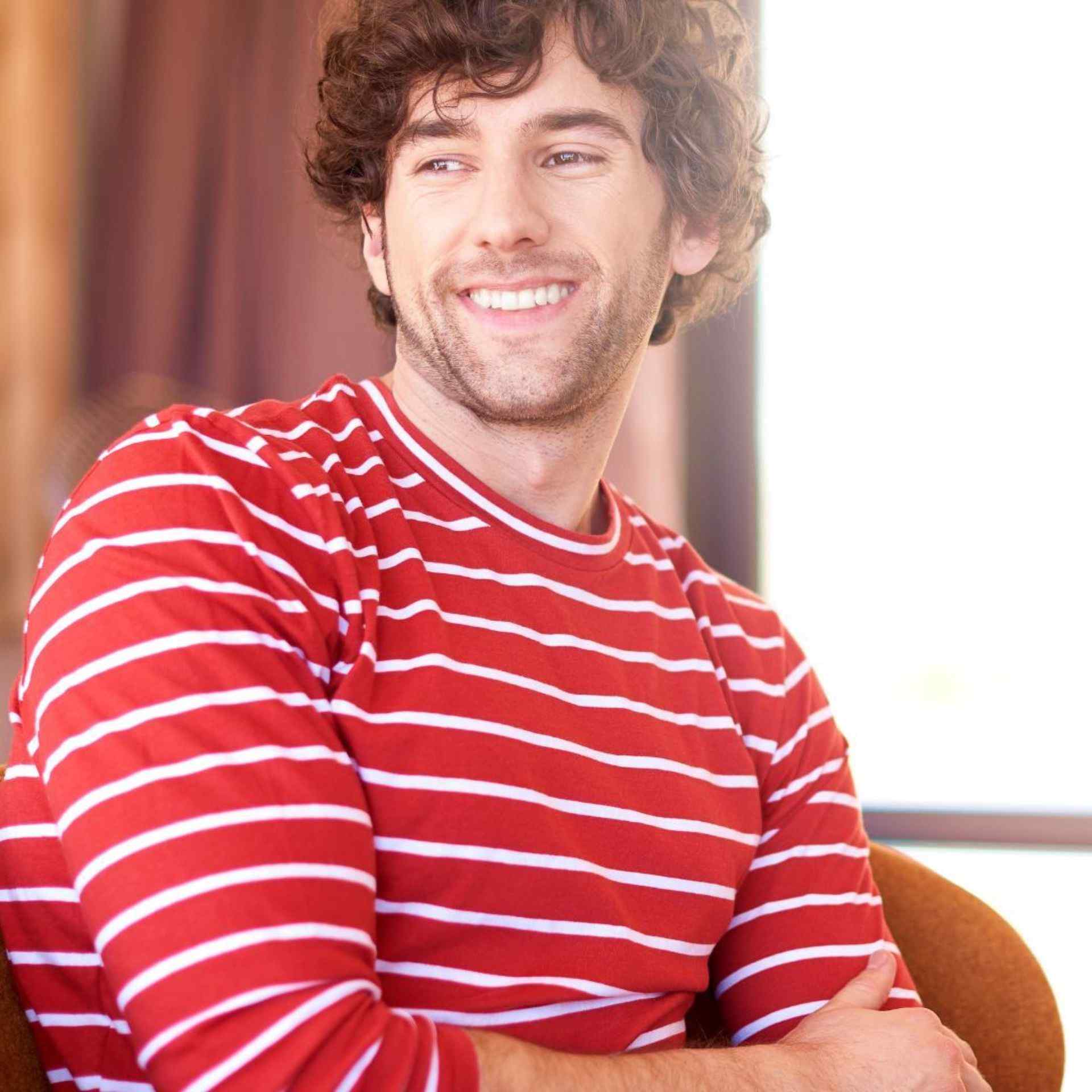 A man with curly hair smiles warmly, wearing a red and white striped shirt. He sits casually with arms crossed, exuding a relaxed and cheerful vibe.