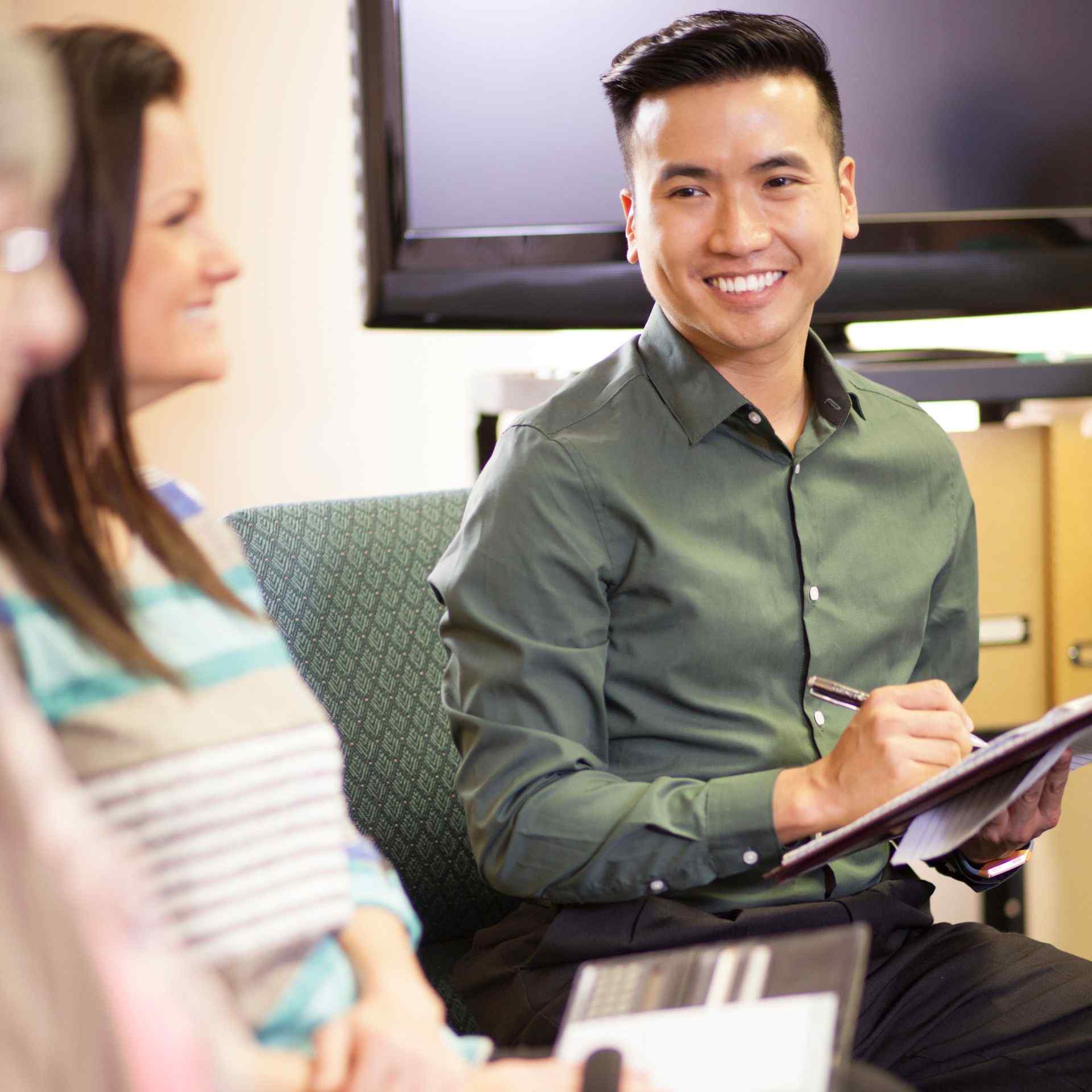 A person in a green shirt smiles while taking notes during a meeting. They are engaged with two others, creating a friendly and collaborative atmosphere.