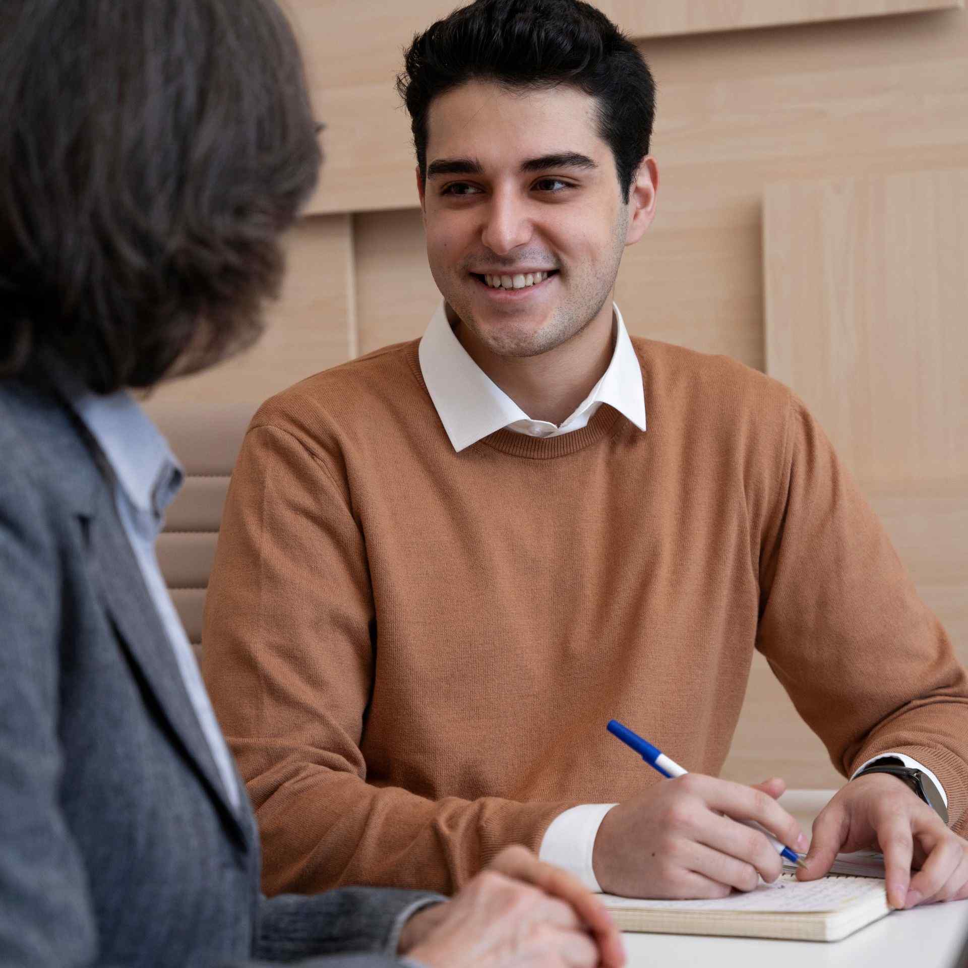 A man in a brown sweater smiles during a conversation with a woman in a suit. He holds a pen over a notebook, suggesting a friendly, professional setting.