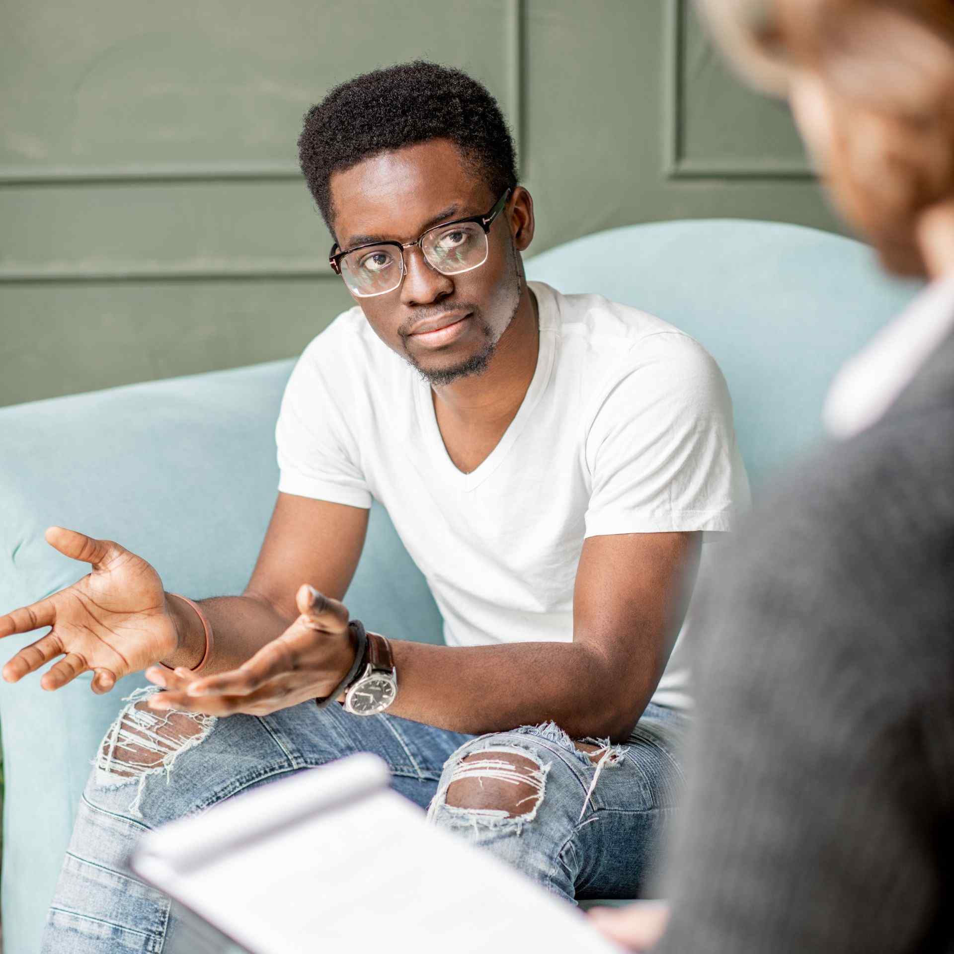 A man in a white t-shirt and ripped jeans sits on a blue couch, gesturing with his hands during a conversation, conveying engagement and focus.