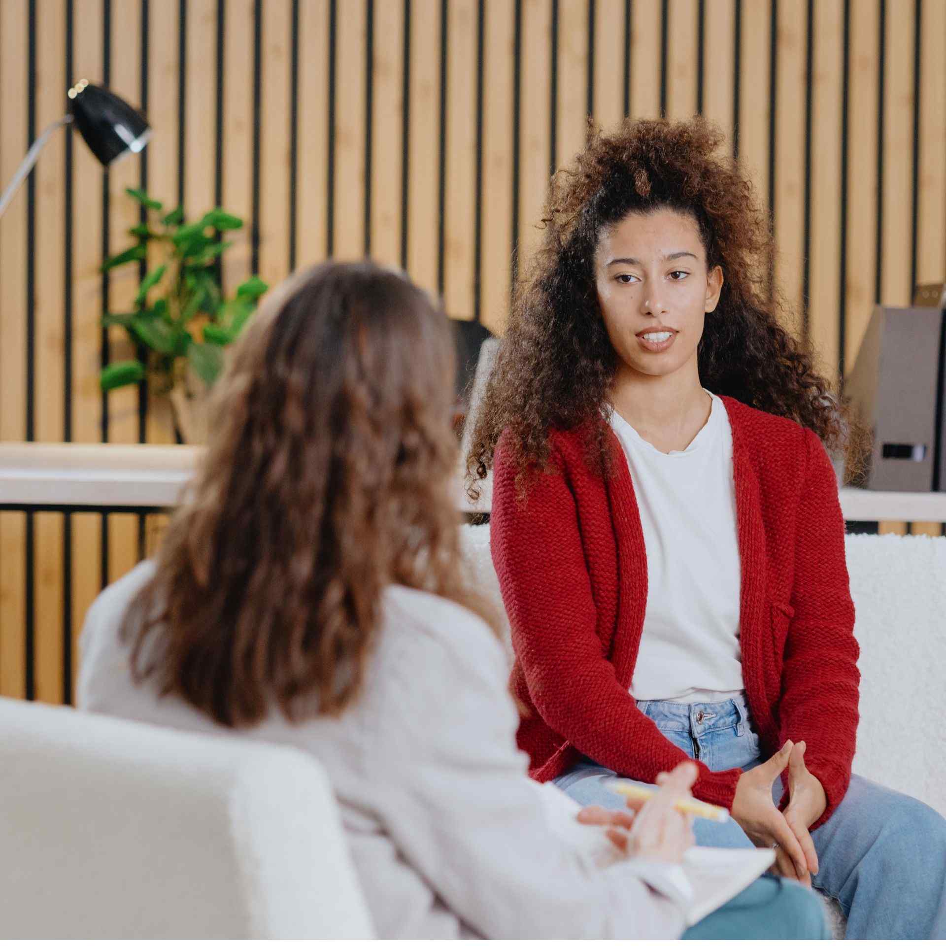 A woman in a red sweater, seated, speaks to another woman with curly hair in an office setting. The mood is focused and engaged. Background features wooden paneling and plants.